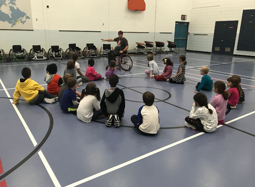 Group of children sitting on a gym floor looking at wheelchairs adapted for sport during an awareness experience school session by NASA.